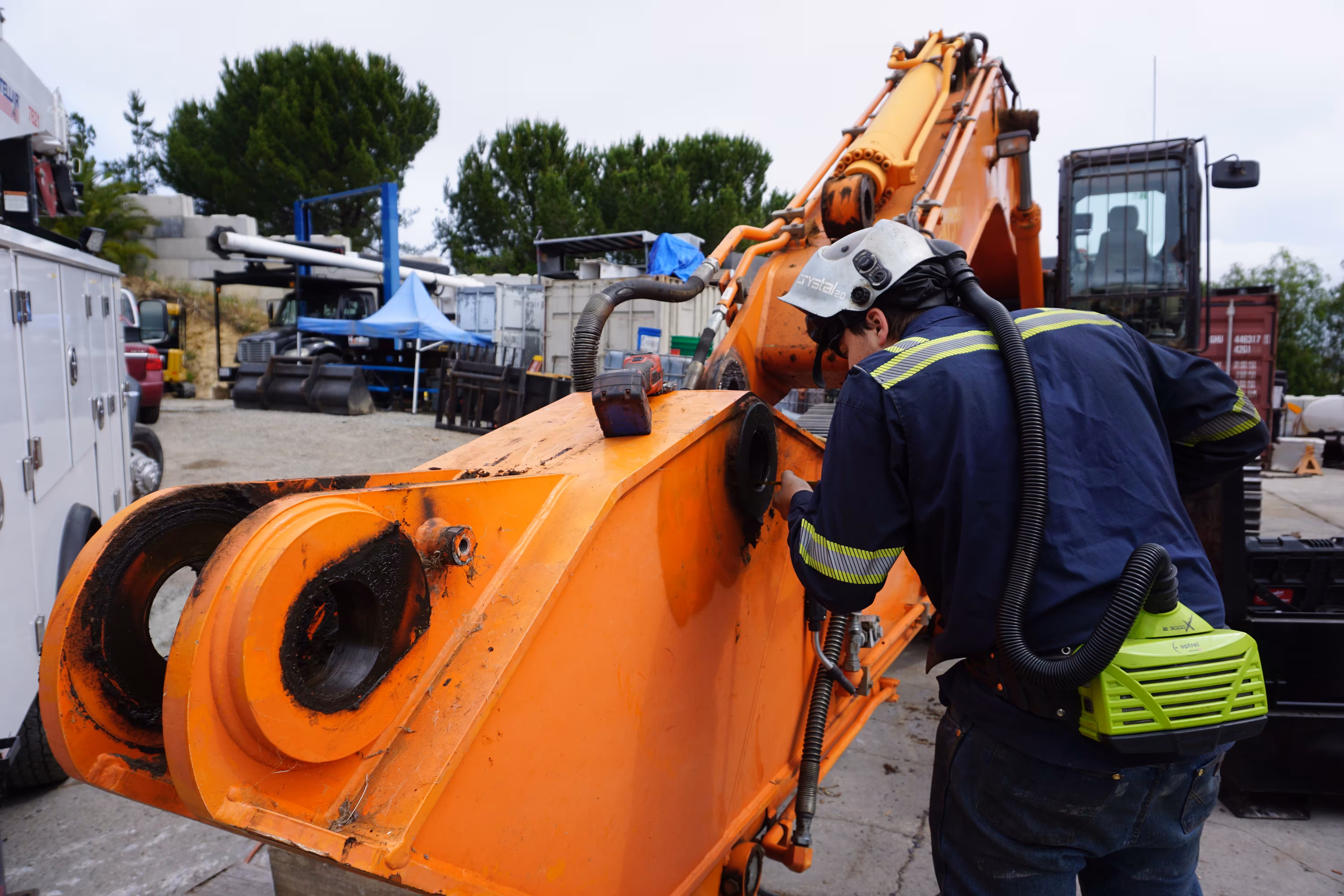 A technician in a respirator hood works on the worn pin bores of an orange excavator boom