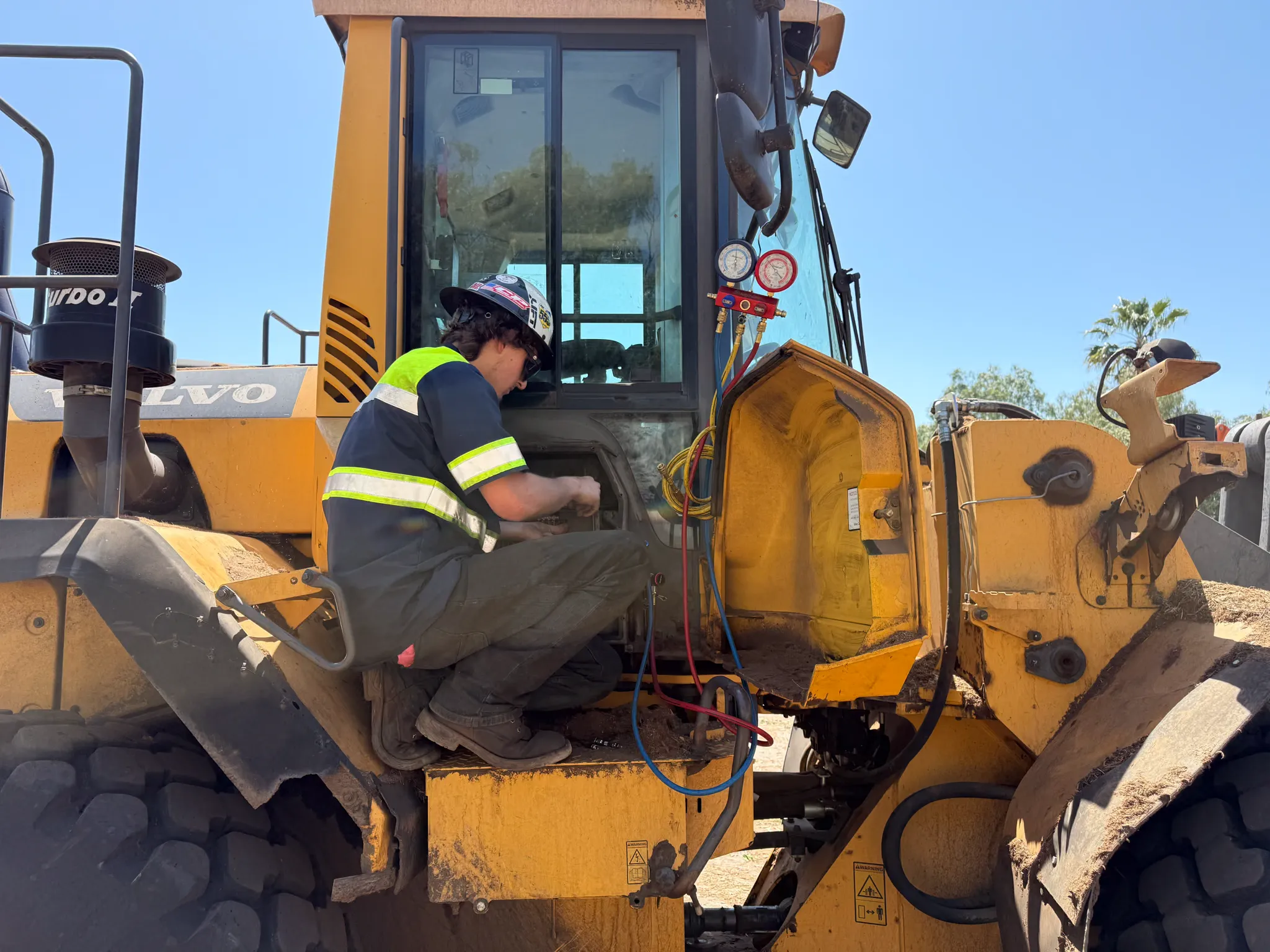 A technician connects A/C manifold gauges to a Volvo wheel loader at a dirt jobsite
