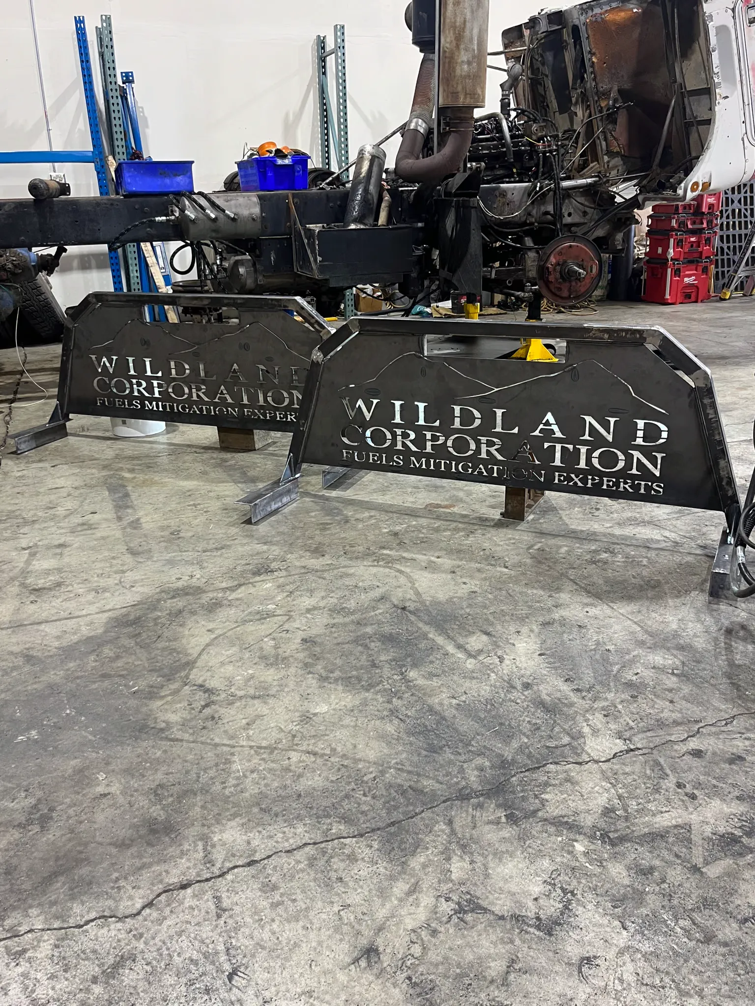 Two custom-cut steel headache racks on the shop floor beside a stripped truck chassis