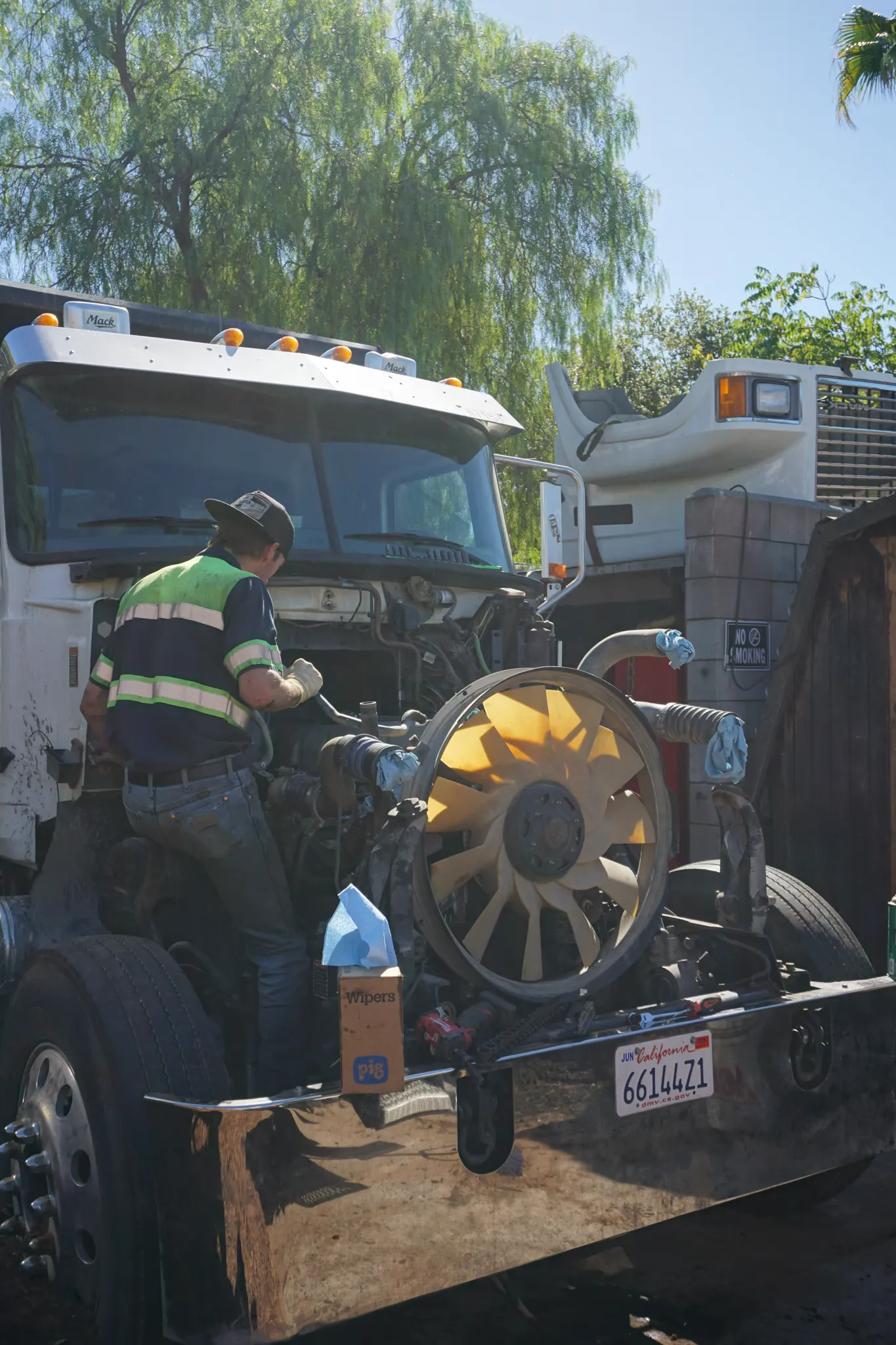 A technician works in the open engine bay of a Mack dump truck, cooling fan exposed