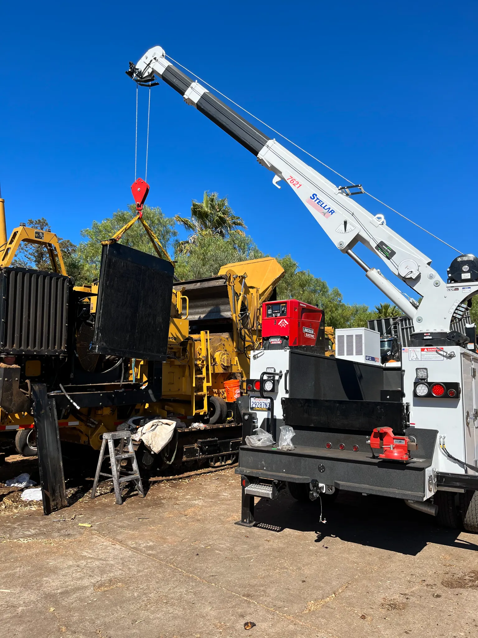 A truck-mounted Stellar crane lifts a steel panel off a yellow tracked grinder in a work yard