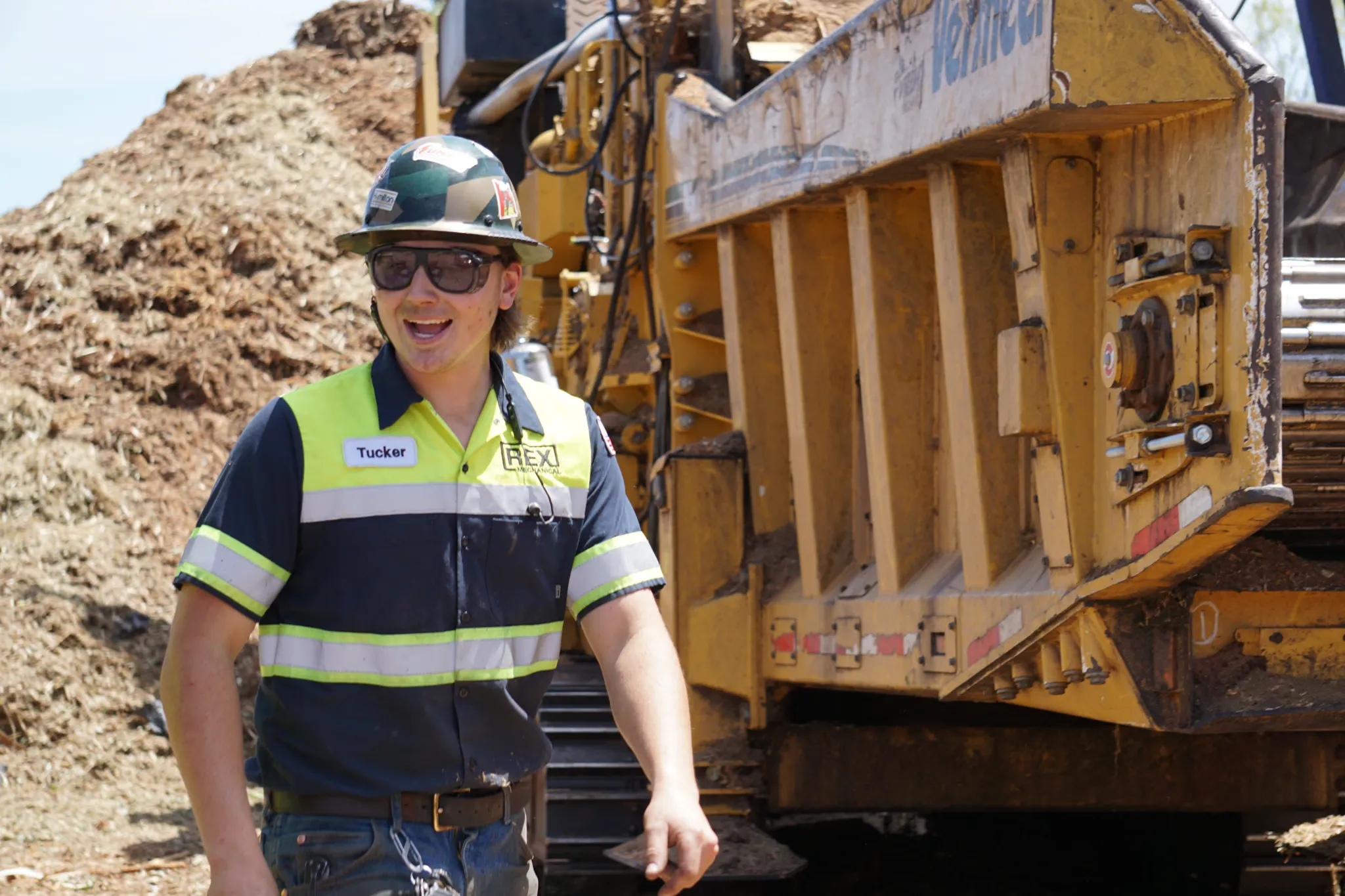 Technician in a REX Mechanical shirt beside a tracked Vermeer grinder next to a mulch pile