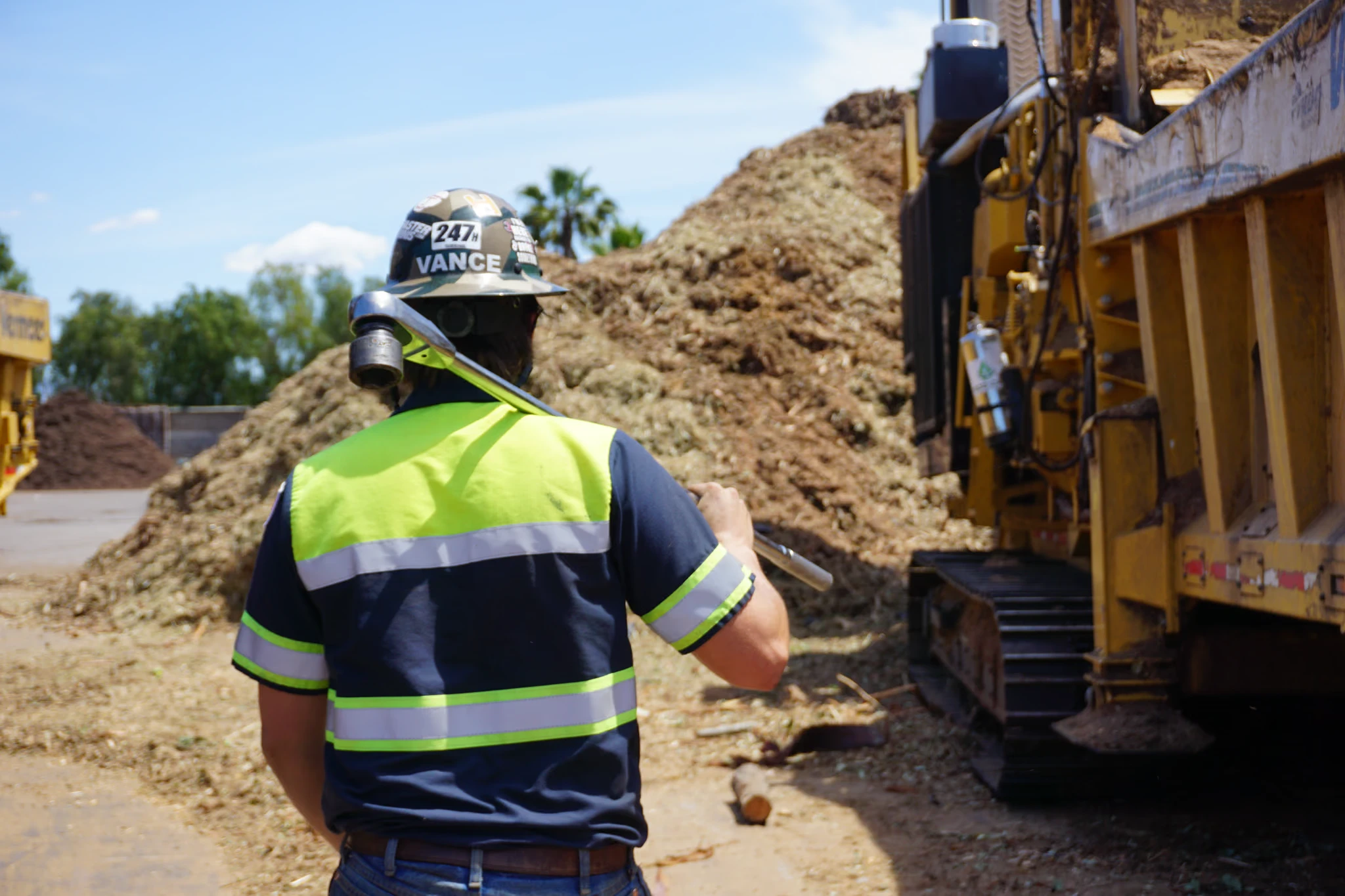 A technician carries a long wrench toward a yellow tracked machine beside mulch piles in a yard
