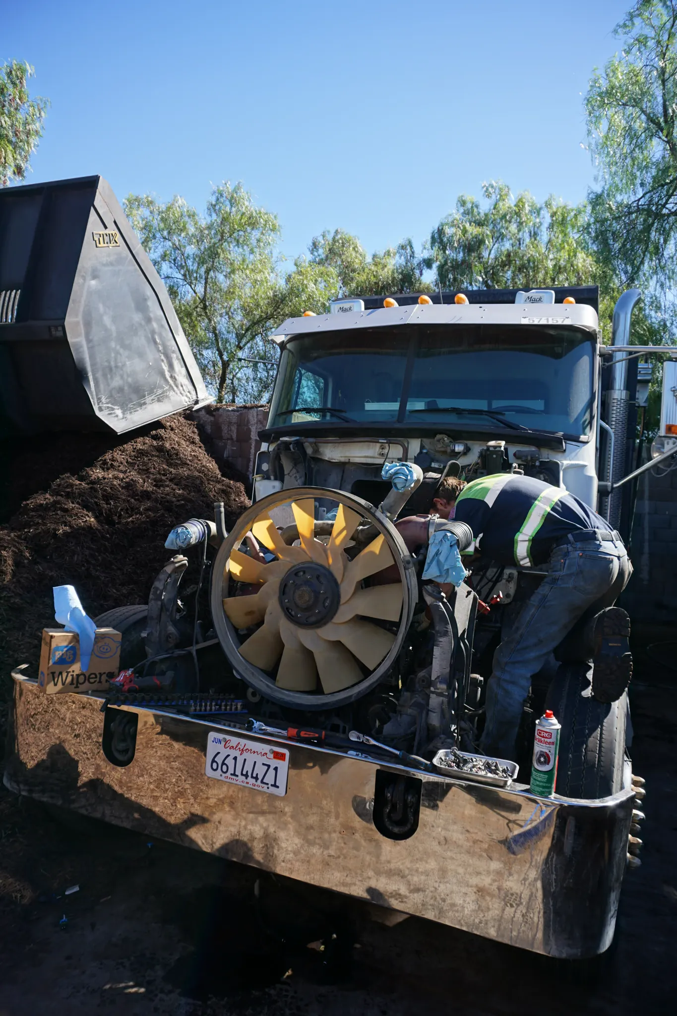 A technician works in the open engine bay of a Mack truck at a mulch yard, cooling fan exposed