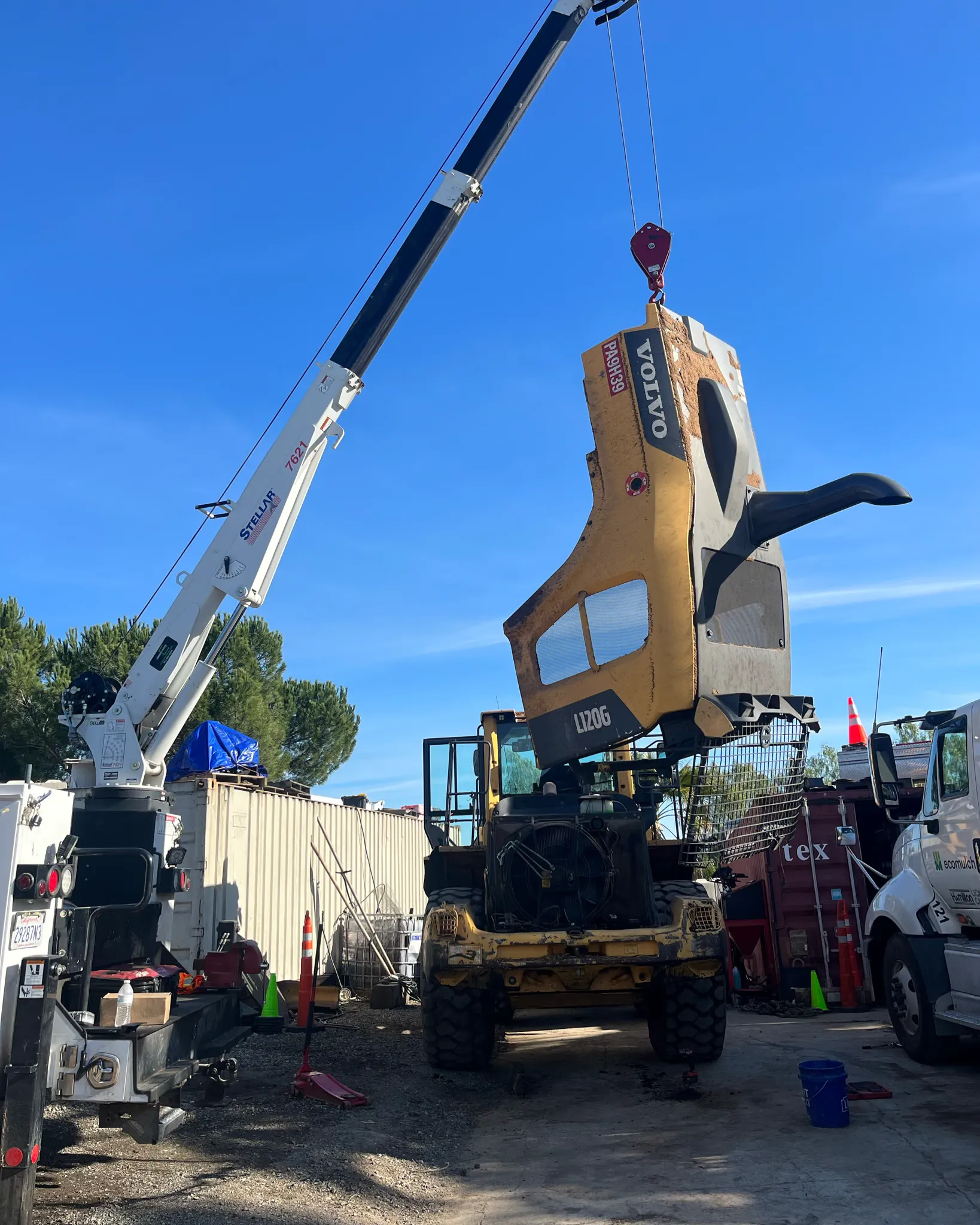 A truck-mounted crane lifts the engine hood off a Volvo L120G wheel loader in the yard