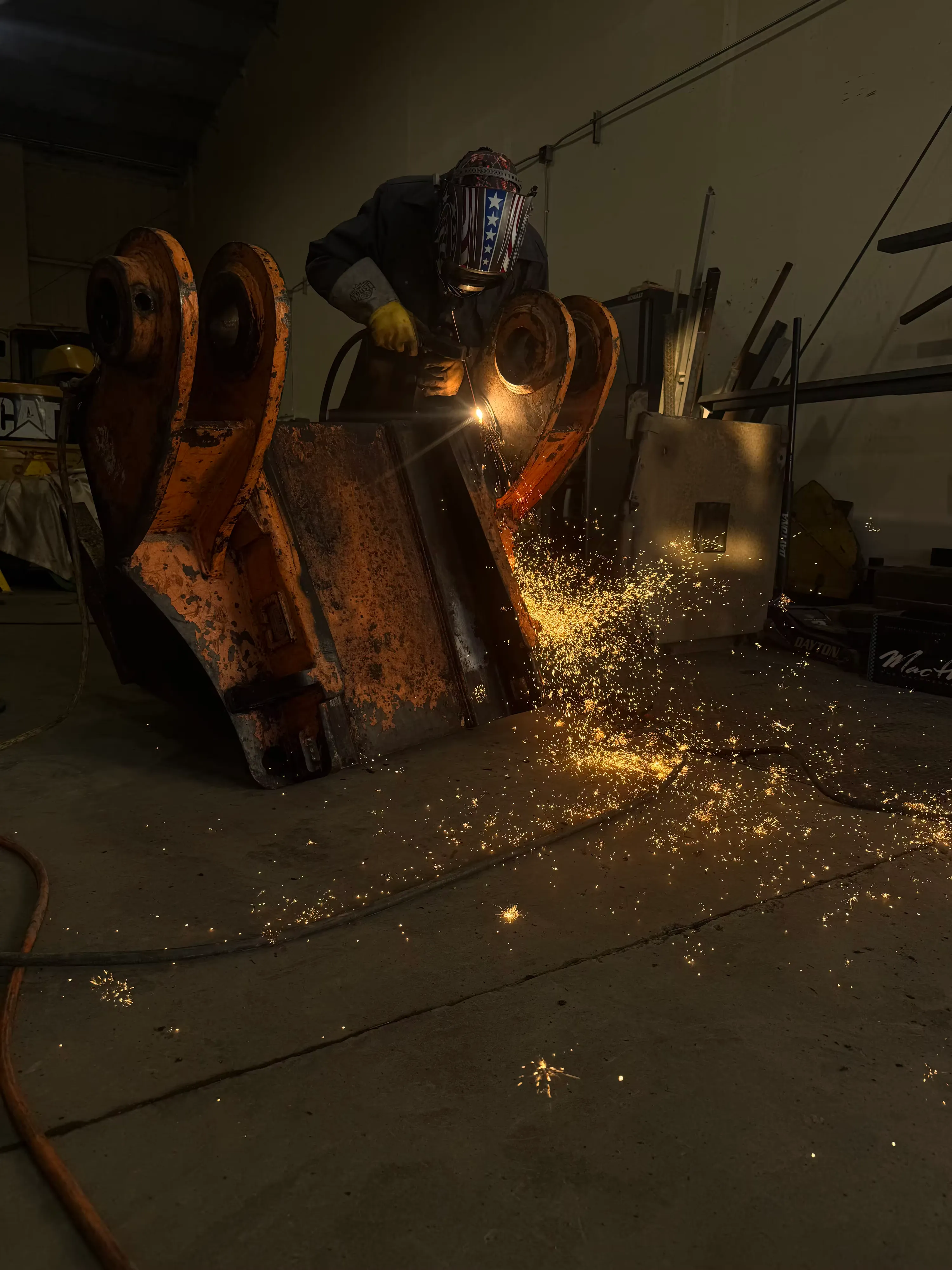 A technician welds a pin boss on a large excavator bucket, sparks arcing across the shop floor